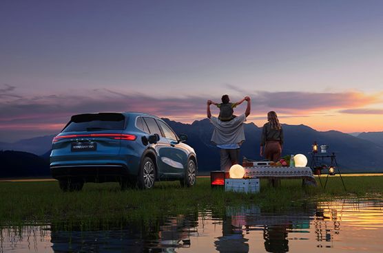 Geely Starray EM-i bei Sonnenuntergang vor einer Bergkulisse am Wasser, im Vordergrund ein Vater mit Kind auf den Schultern und eine Frau bei einem Campingaufbau mit Lichtern und Picknick.