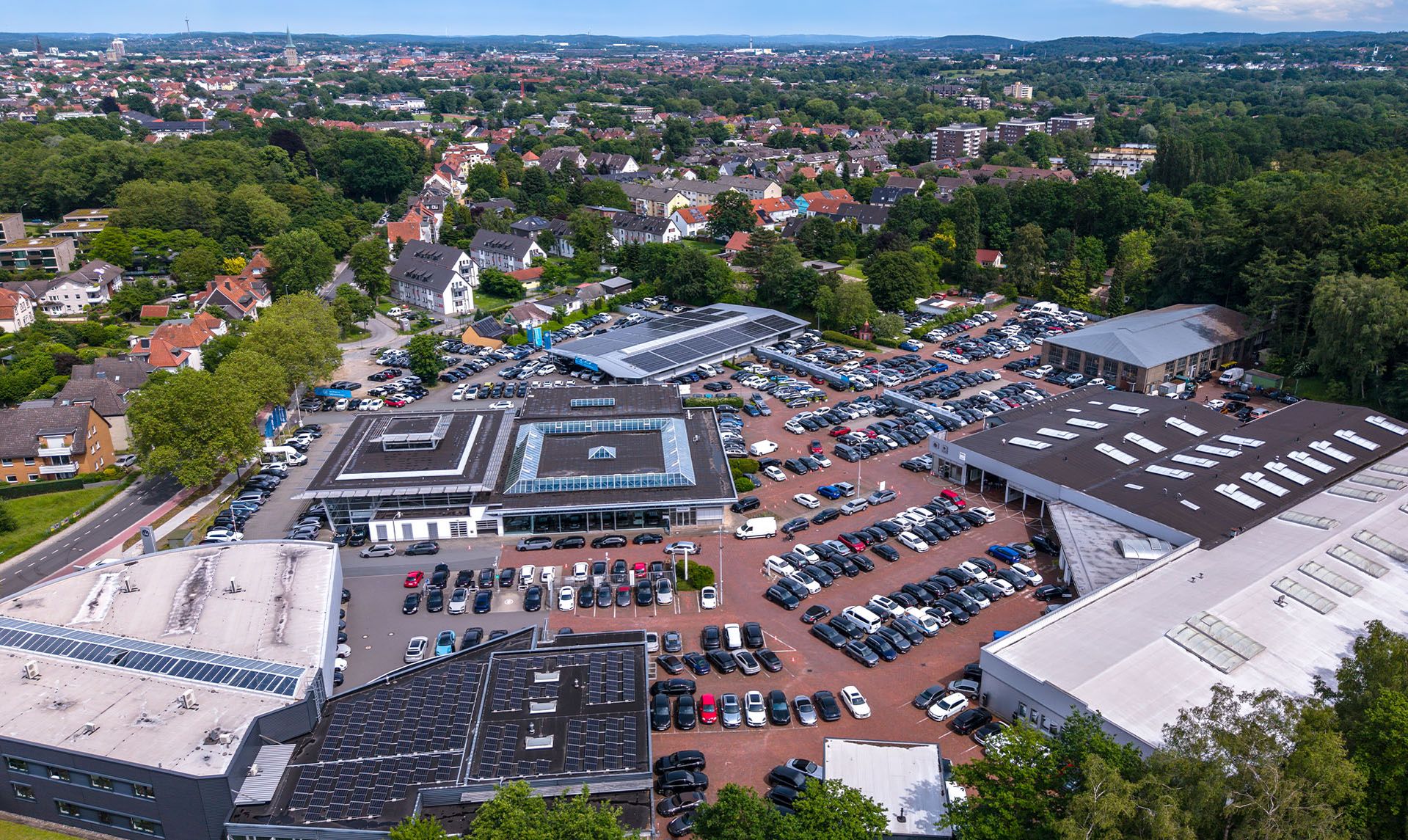 Luftaufnahme eines Automobilstandorts mit Ausstellungsflächen, Servicegebäuden und zahlreichen Fahrzeugen vor einer städtischen Umgebung.