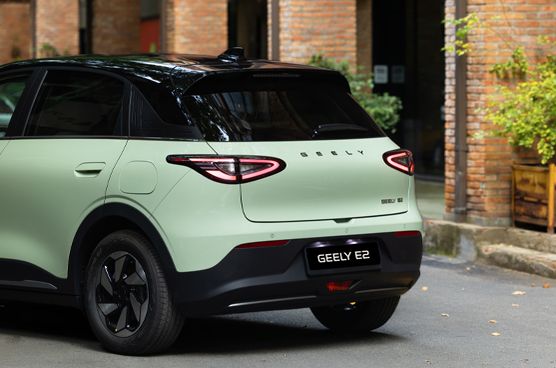 Geely E2 in Heckseitenansicht auf einer Straße vor einem Gebäude mit Backsteinfassade und Pflanzen.
