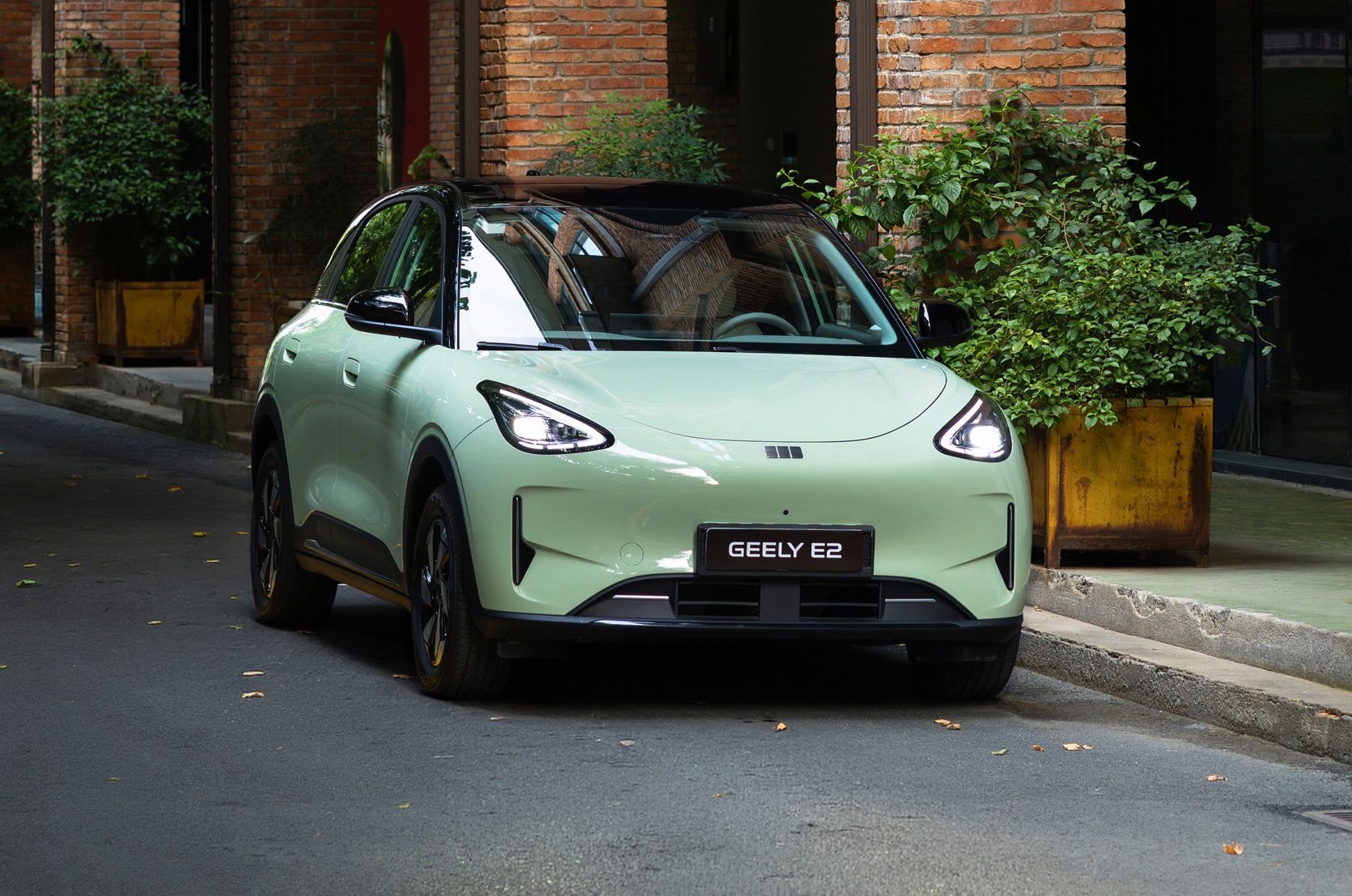 Geely E2 in hellgrüner Lackierung auf einer Straße vor einem Gebäude mit Backsteinfassade und bepflanzten Eingangsbereichen.