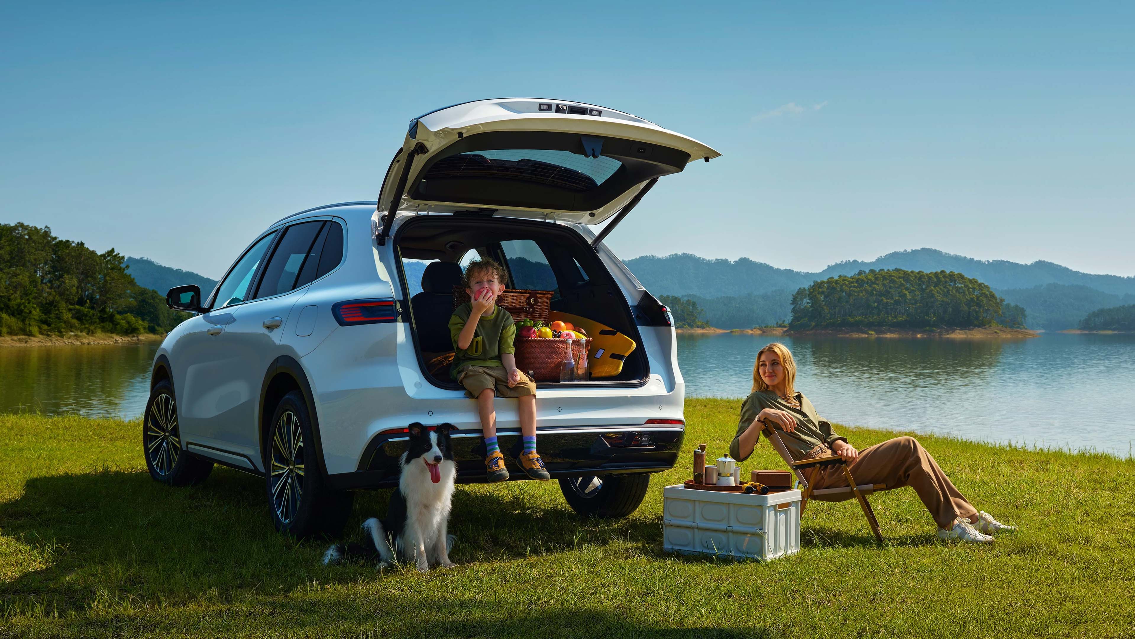 Geely Starray EM-i mit geöffneter Heckklappe und Picknickkorb am Ufer eines Sees, im Vordergrund ein Junge auf der Ladekante und eine Frau auf einem Klappstuhl.