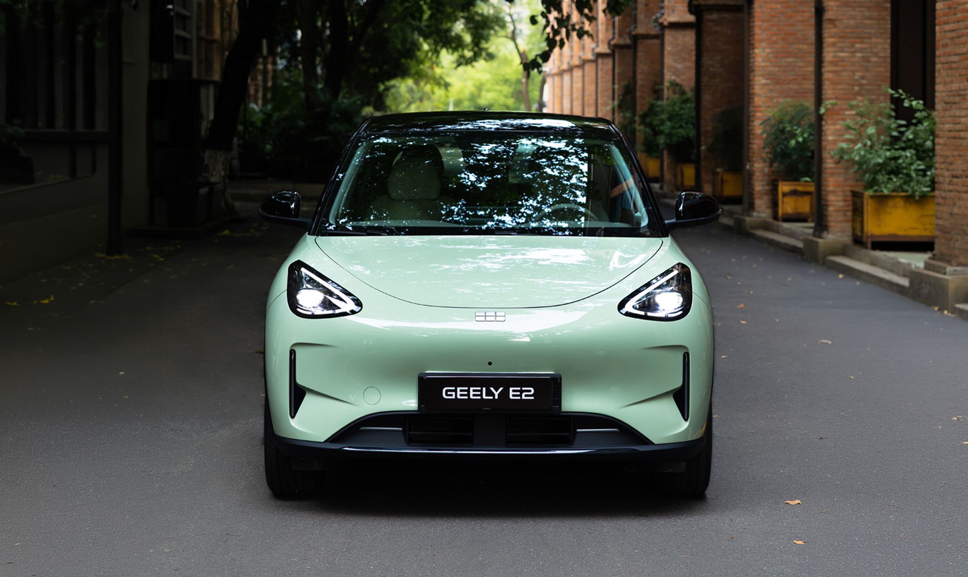 Geely E2 in Frontansicht auf einer Straße zwischen Backsteingebäuden und bepflanzten Bereichen.