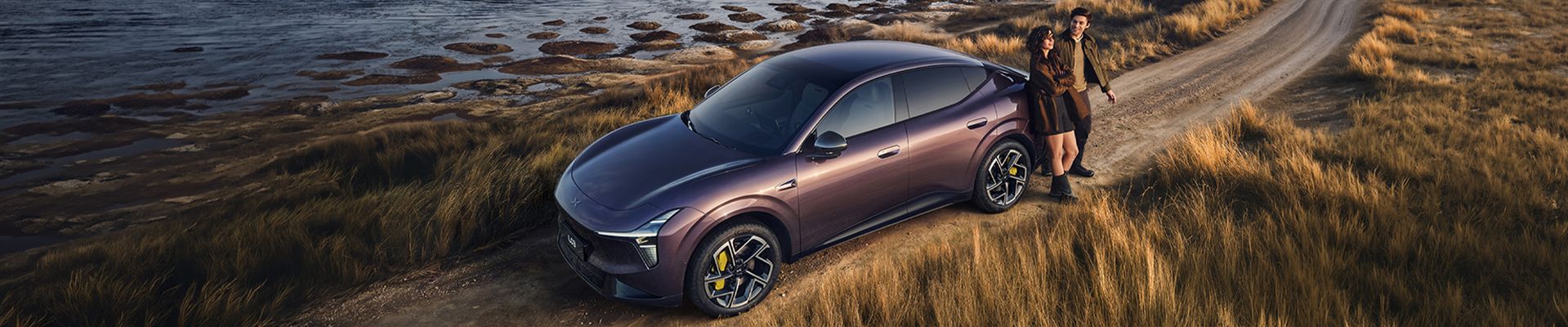 Ein violetter XPENG L03 steht auf einem Feldweg an einer Küstenlandschaft im Abendlicht. Neben dem SUV-Coupé steht ein Paar und blickt auf das Wasser.