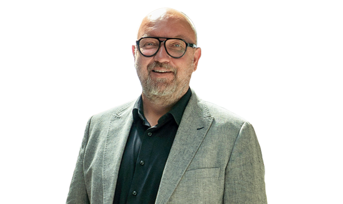 Ein älterer Mann trägt Brille und schwarzes Hemd mit einem hellen Sakko.