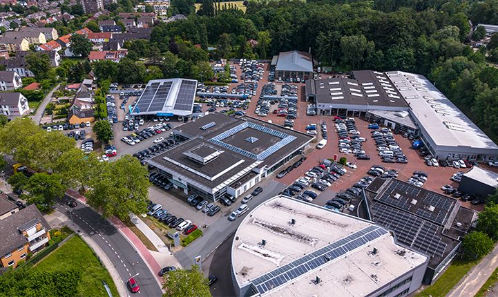 Luftaufnahme VW Zentrum mit mehreren Ausstellungsflächen, Fahrzeugstellplätzen und Gebäuden in einer von Bäumen umgebenen Umgebung.