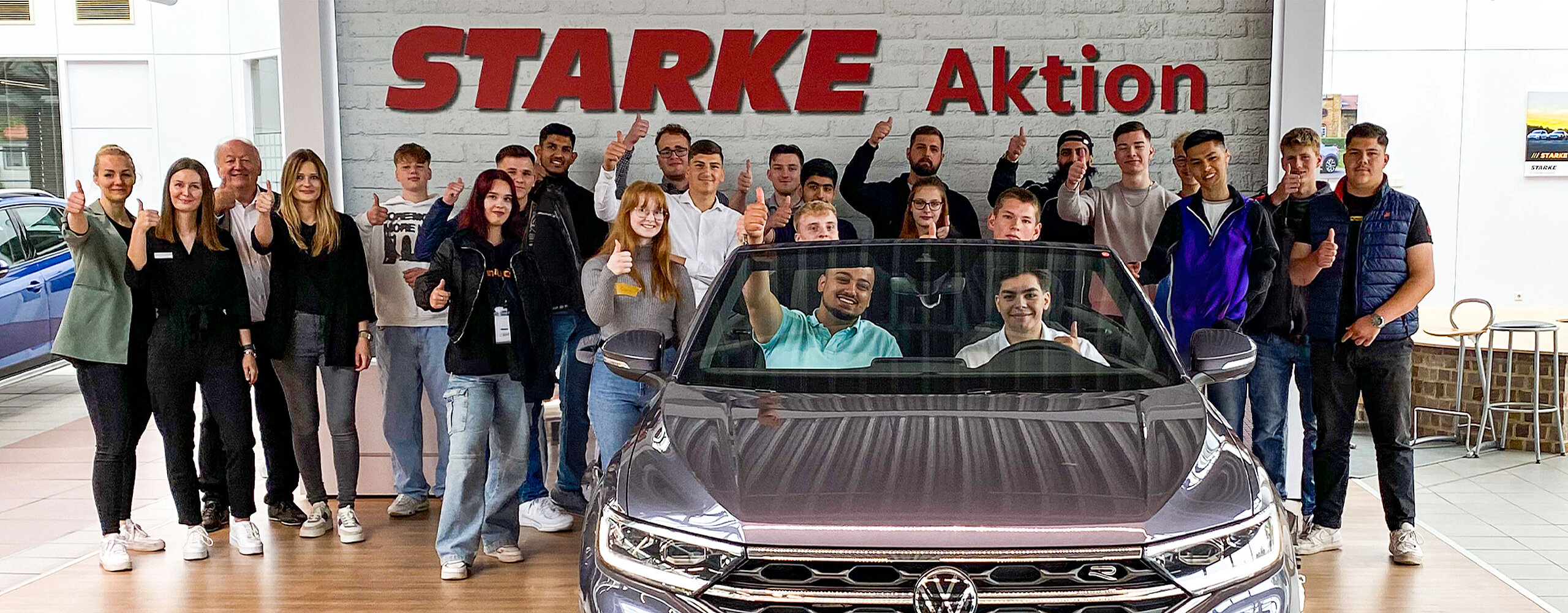 Dieses Bild zeigt ein Gruppenbild von allen Auszubildenden der STARKE Gruppe. Zwei männliche Azubis sitzen in einem VW Cabrio. Im Hintergrund an der Wand steht der Slogan: STARKE Aktion