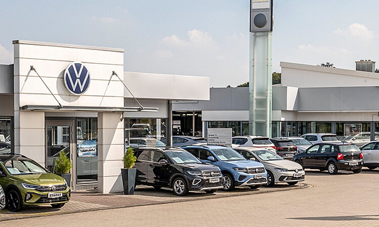 Auf dem Bild ist der Volkswagen Standort in Georgsmarienhütte zu sehen. Im Vordergrund steht der Eingang mit einem Volkswagen Logo. Im Hintergrund ist der geräumige Parkplatz zu sehen. 