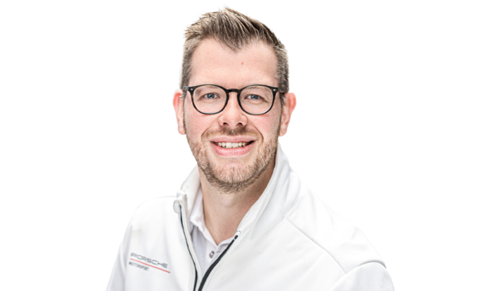 Mitarbeiter in weißer Porsche Jacke und Brille blickt lächelnd in die Kamera. Im Hintergrund sind unscharf Fahrzeuge in einem Autohaus zu erkennen.