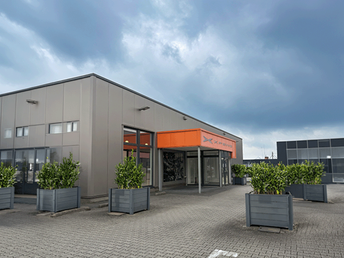 Eingangsbereich eines XPENG Showrooms mit orangefarbener Fassadenverkleidung, XPENG Markenlogo und bepflanzten Kübeln.