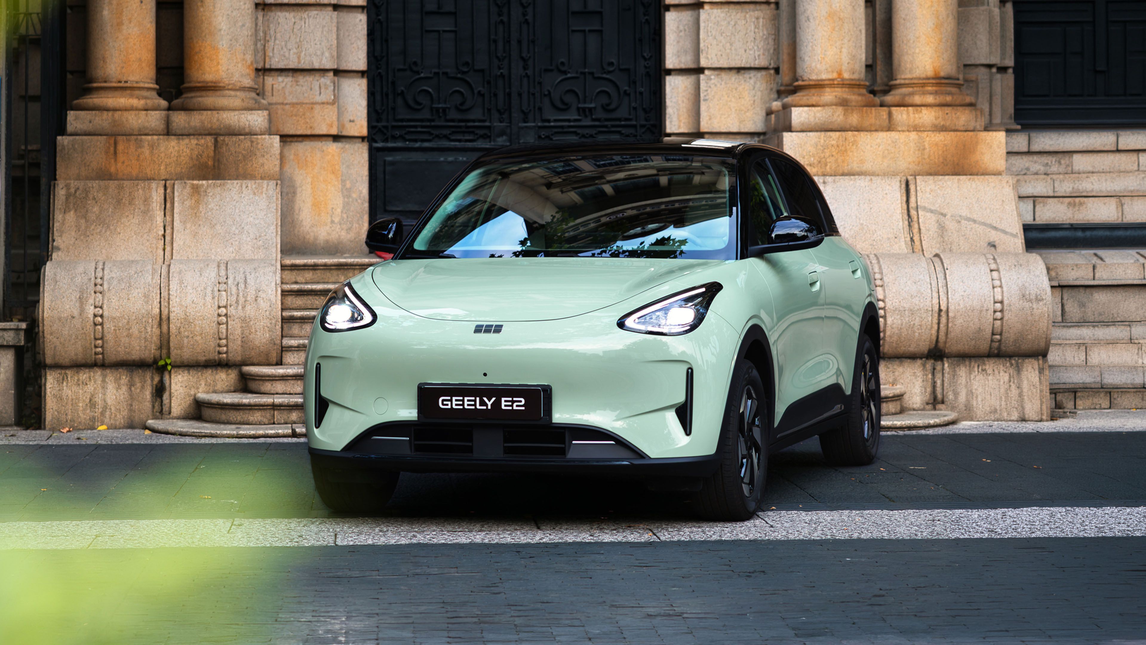 Geely E2 in hellgrüner Lackierung vor einem Gebäude mit Natursteinfassade und Säulen.