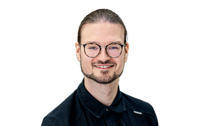 Portrait eines Mitarbeiters mit zurückgegeeltem Haar, Bart und Brille in einem schwarzen Hemd.