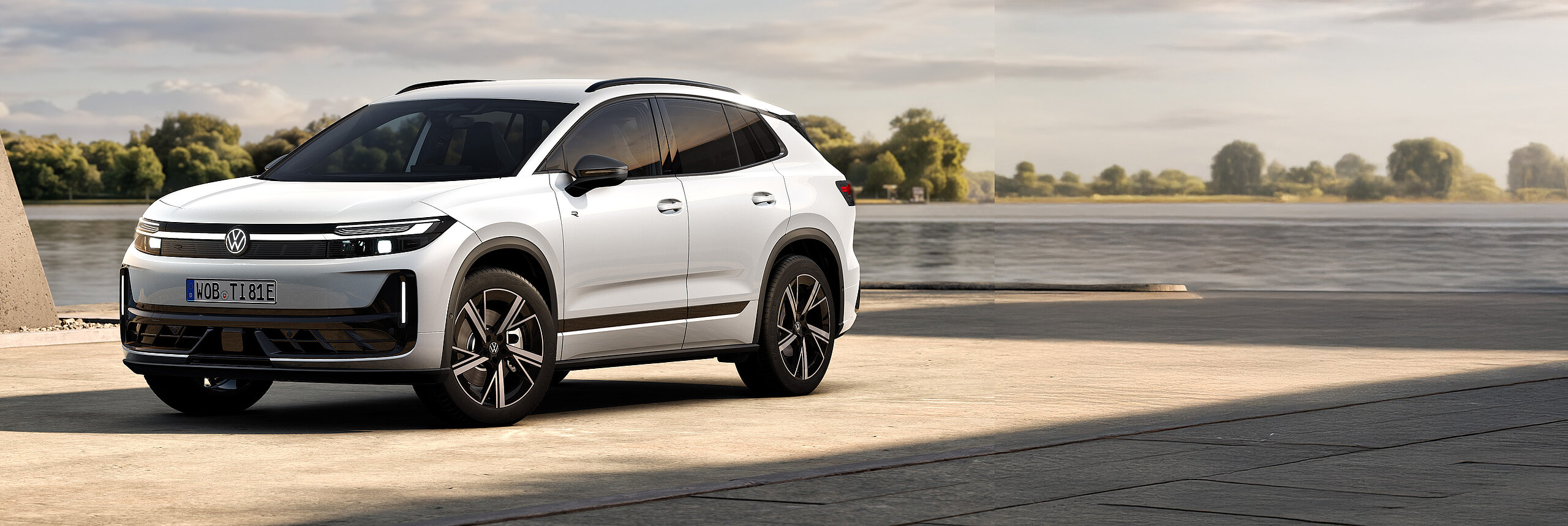 Volkswagen ID. Tiguan in weißer Lackierung in Frontseitenansicht auf einer befestigten Fläche vor einem Gewässer mit Bäumen am Ufer.