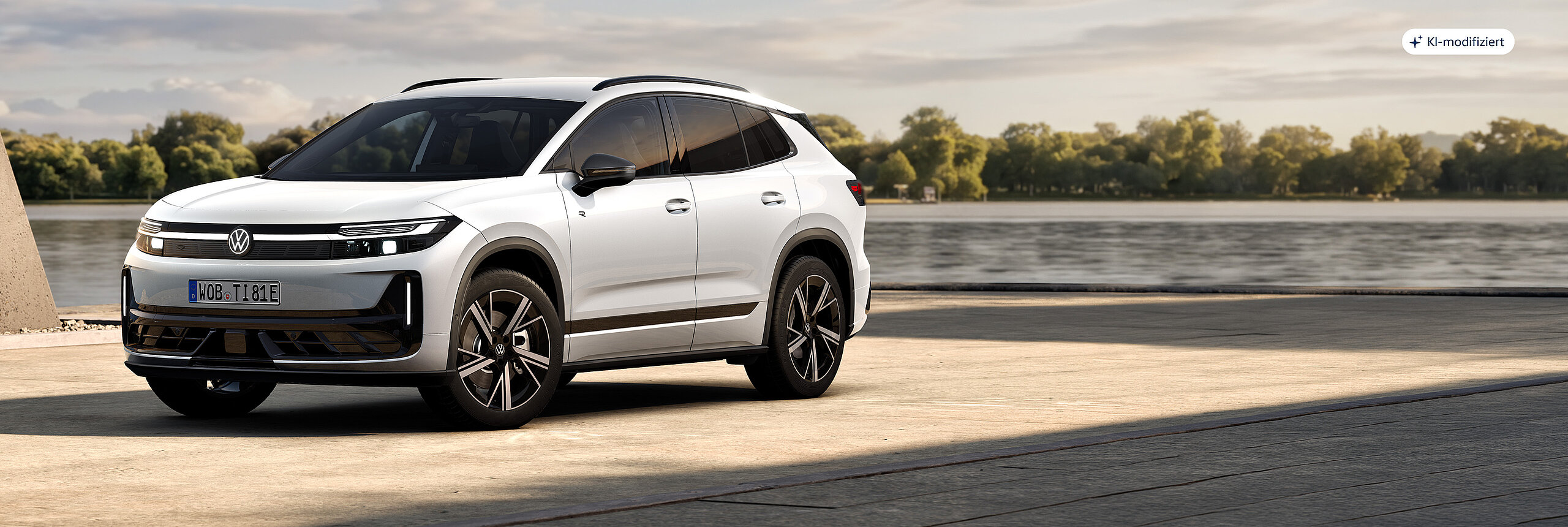 Volkswagen ID. Tiguan in weißer Lackierung in Frontseitenansicht auf einer befestigten Fläche vor einem Gewässer mit Bäumen am Ufer.