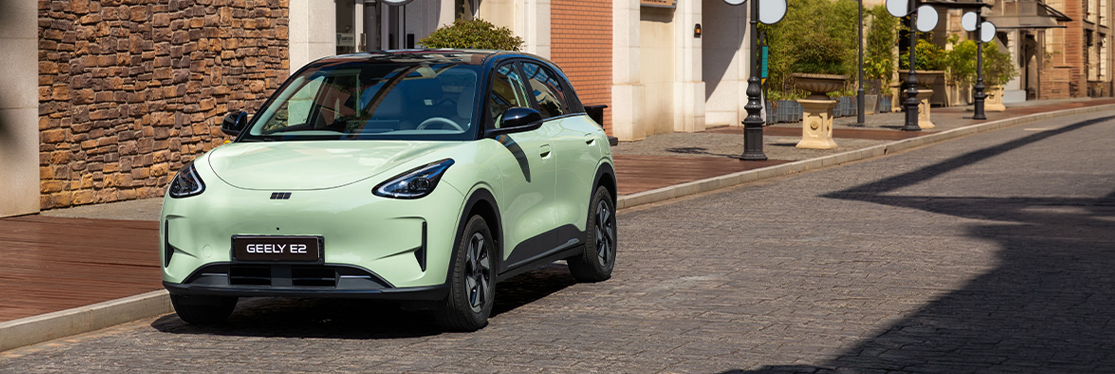 Geely E2 in hellgrüner Lackierung auf einer gepflasterten Straße vor Gebäuden mit Stein- und Backsteinfassaden.