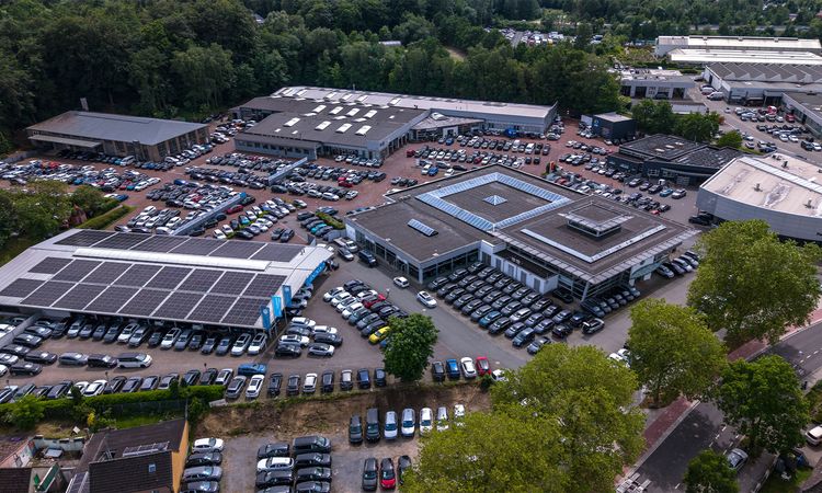 Luftaufnahme eines Automobilstandorts mit Ausstellungsgebäuden, Fahrzeugstellplätzen und zahlreichen Fahrzeugen in einer von Bäumen umgebenen Umgebung.