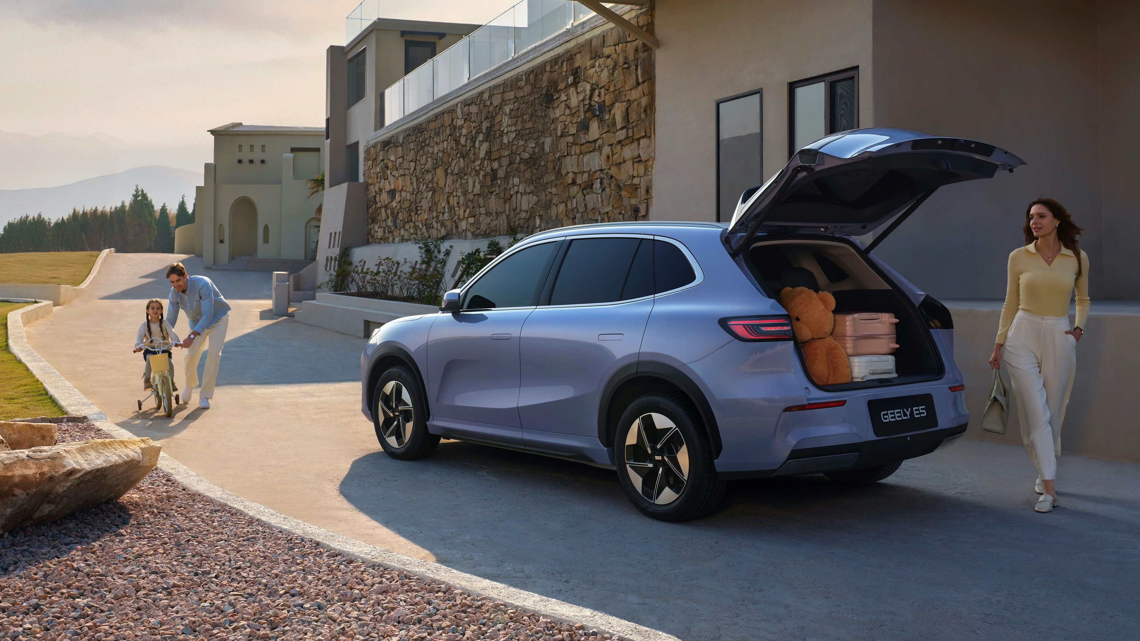 Geely E5 mit geöffneter Heckklappe und Gepäck vor einem modernen Wohnhaus, daneben Familie mit Kind auf dem Fahrrad.