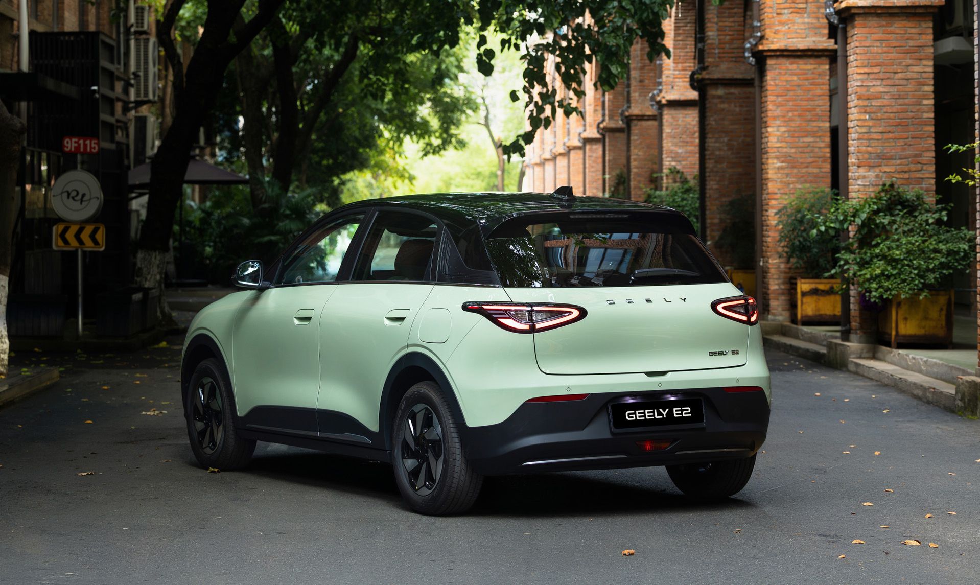 Geely E2 in Heckseitenansicht auf einer Straße mit Bäumen und Backsteingebäuden im Hintergrund.