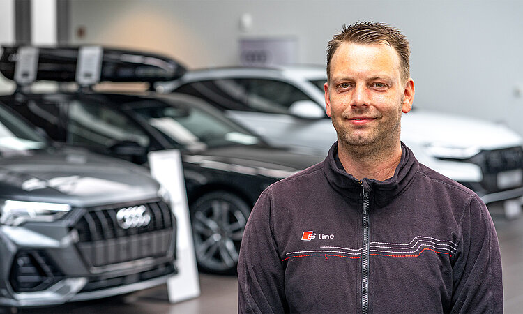 Auf dem Bild ist im Vordergrund ein Mann abgebildet, er hat kurze dunkelblonde Haare und lächelt in die Kamera. Im Hintergrund sit etwas verschwommen der Audi Verkaufsraum des Autohauses zu sehen..