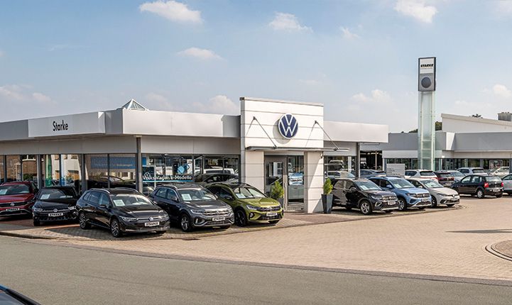 Auf dem Bild ist der Volkswagen Standort in Georgsmarienhütte zu sehen. Im Vordergrund steht der Eingang mit einem Volkswagen Logo. Im Hintergrund ist der geräumige Parkplatz zu sehen. 