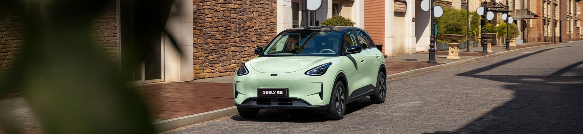 Geely E2 in hellgrüner Lackierung auf einer gepflasterten Straße in einer städtischen Umgebung mit Stein- und Backsteinfassaden.