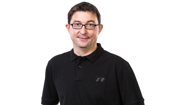 Portrait eines Mitarbeiters mit kurzem Haar und Brille in einem schwarzen Poloshirt.
