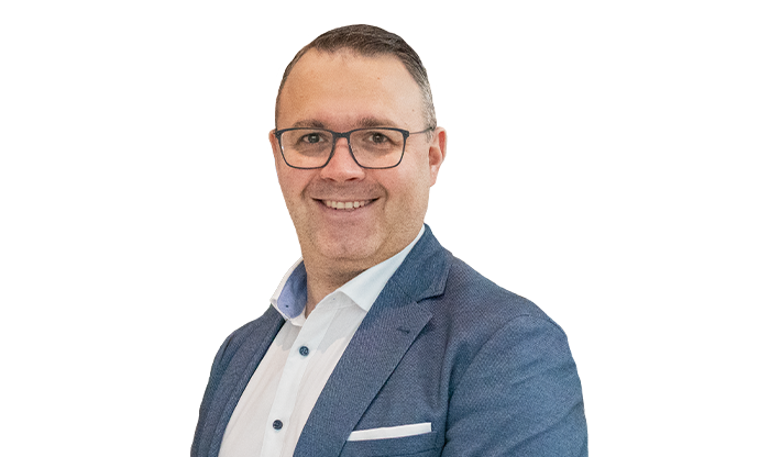 Portrait eines Mitarbeiters mit kurzem Haar und Brille in einem weißen Hemd und einem hellblauen Jackett.
