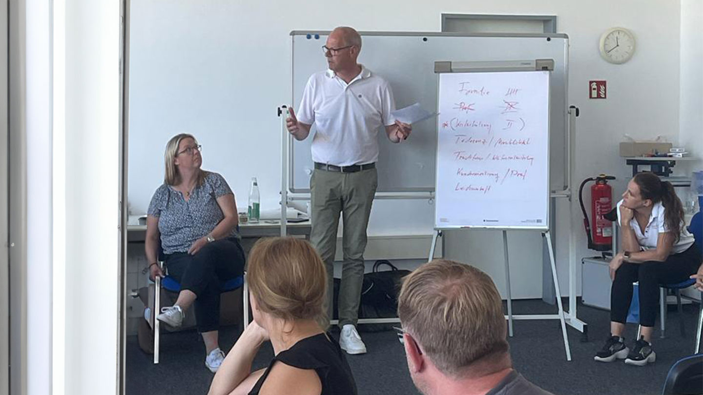 Auf diesem Bild sieht man einen Mann mit Brille und weißem Hemd, der vor einem Flipchart steht und vier Mitarbeitende etwas erklärt.