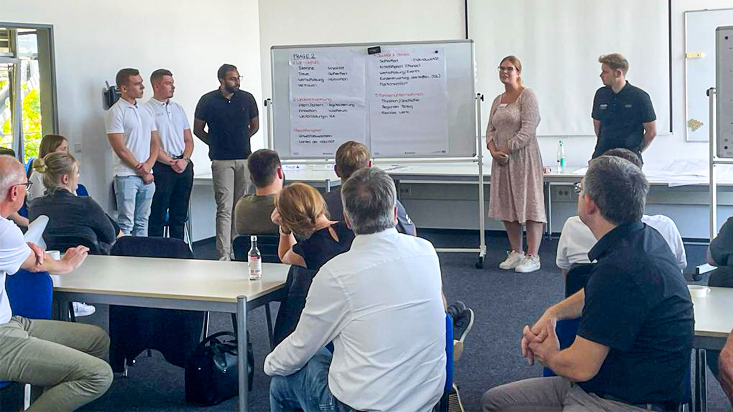 Auf diesem Bild sieht man eine Frau mit Brille und geblümtem Kleid, die neben einem Flipchart steht und mehreren Mitarbeitenden etwas erklärt.