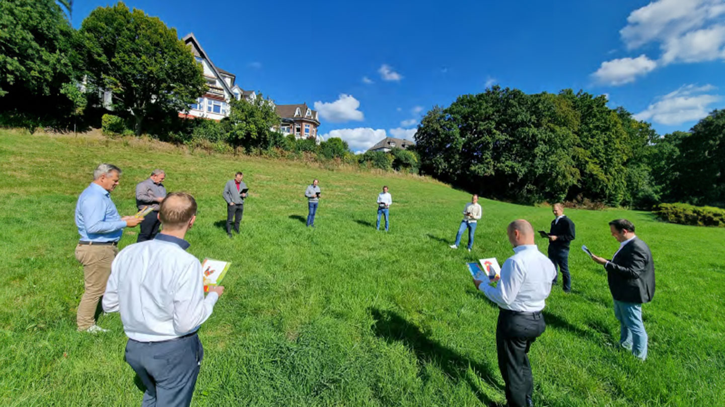 Auf diesem Bild sieht man zehn Männer, die im Kreis auf einer Wiese stehen und jeweils einen Zettel mit einem bunten Bild in der Hand halten.  