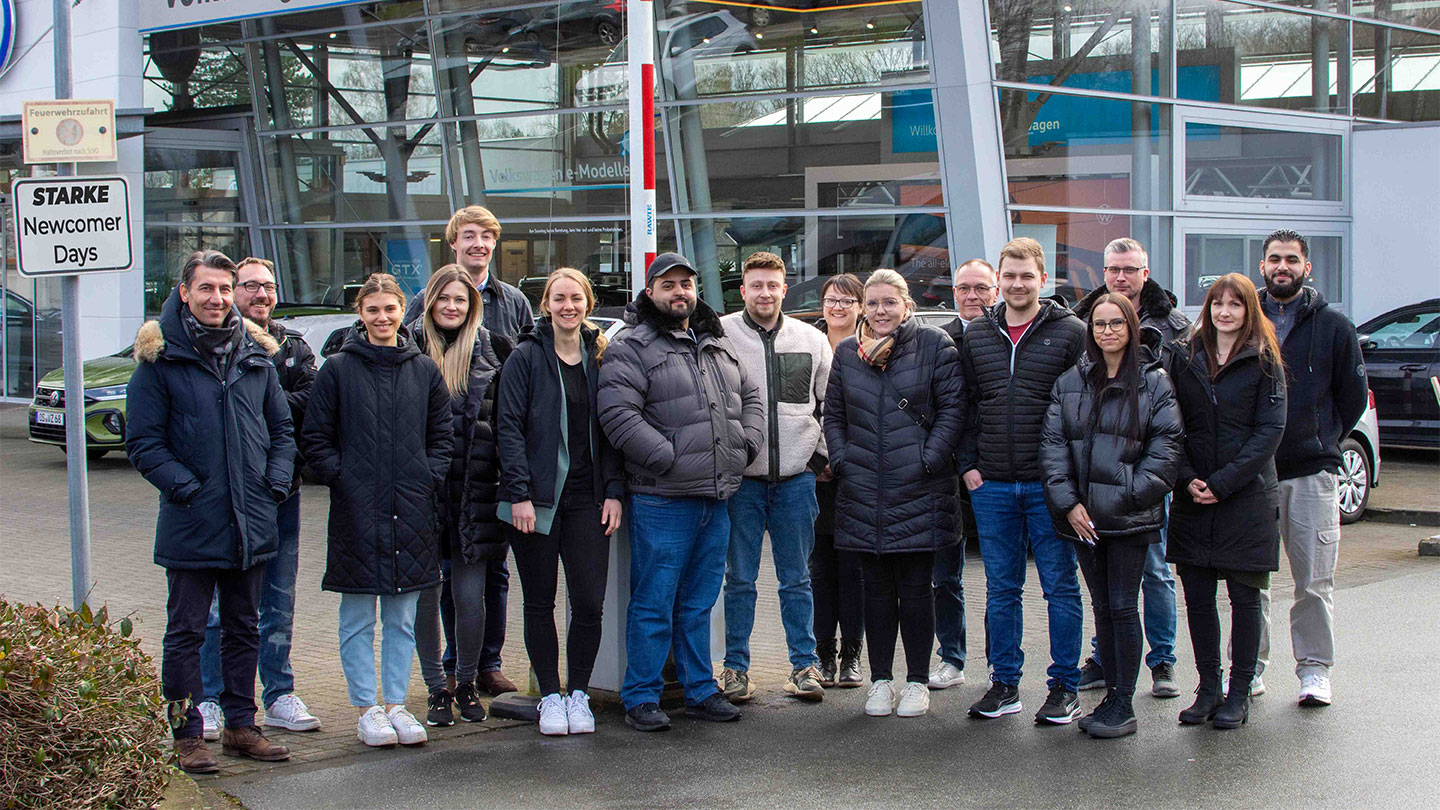 Das Bild zeigt eine Gruppe von Newcomern, die zusammen abgebildet sind, während im Hintergrund ein Autohaus zu sehen ist. 