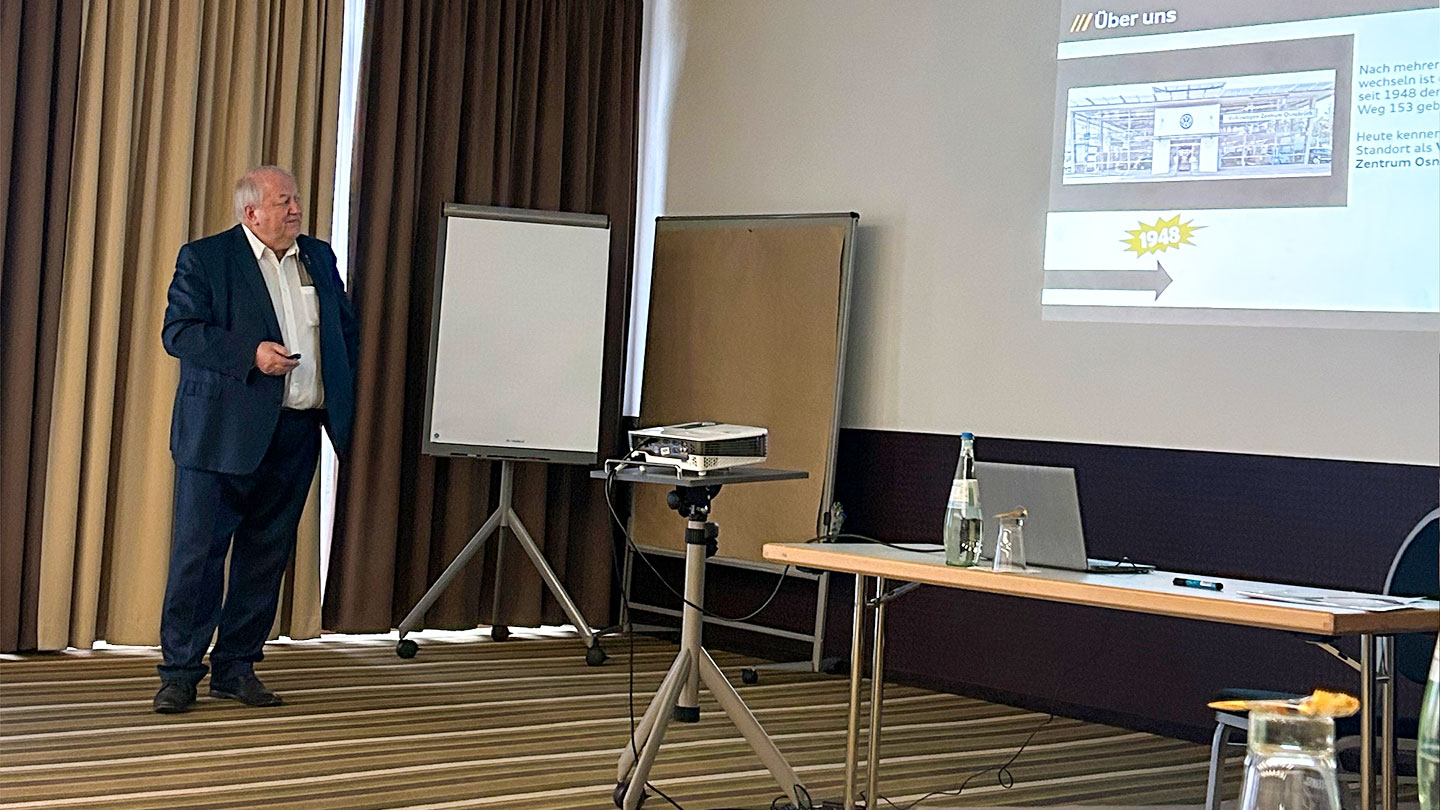 Das Bild zeigt einen Mann in einem weißen Hemd und einem dunklen Sakko, der eine Präsentation mit einem Beamer hält. Das deutet darauf hin, dass er wahrscheinlich vor einer Gruppe spricht, vielleicht bei einem Meeting, einer Schulung oder einer Veranstaltung.