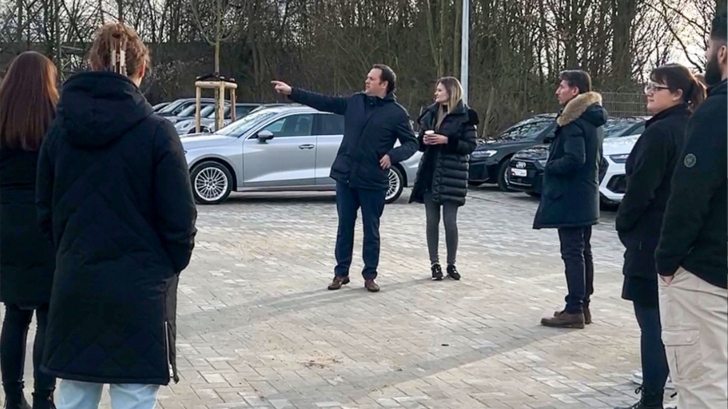 Das Bild zeigt eine Gruppe von Menschen, die auf einem Parkplatz stehen, während ein Mann mit einem Finger in eine Richtung zeigt. Im Hintergrund sind Fahrzeuge zu sehen.