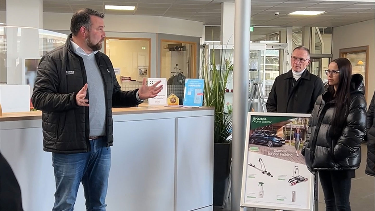 Das Bild zeigt zwei Männer und eine Frau, die sich vor einem Empfangstresen unterhalten. Im Hintergrund ist das Autohaus zu sehen. 