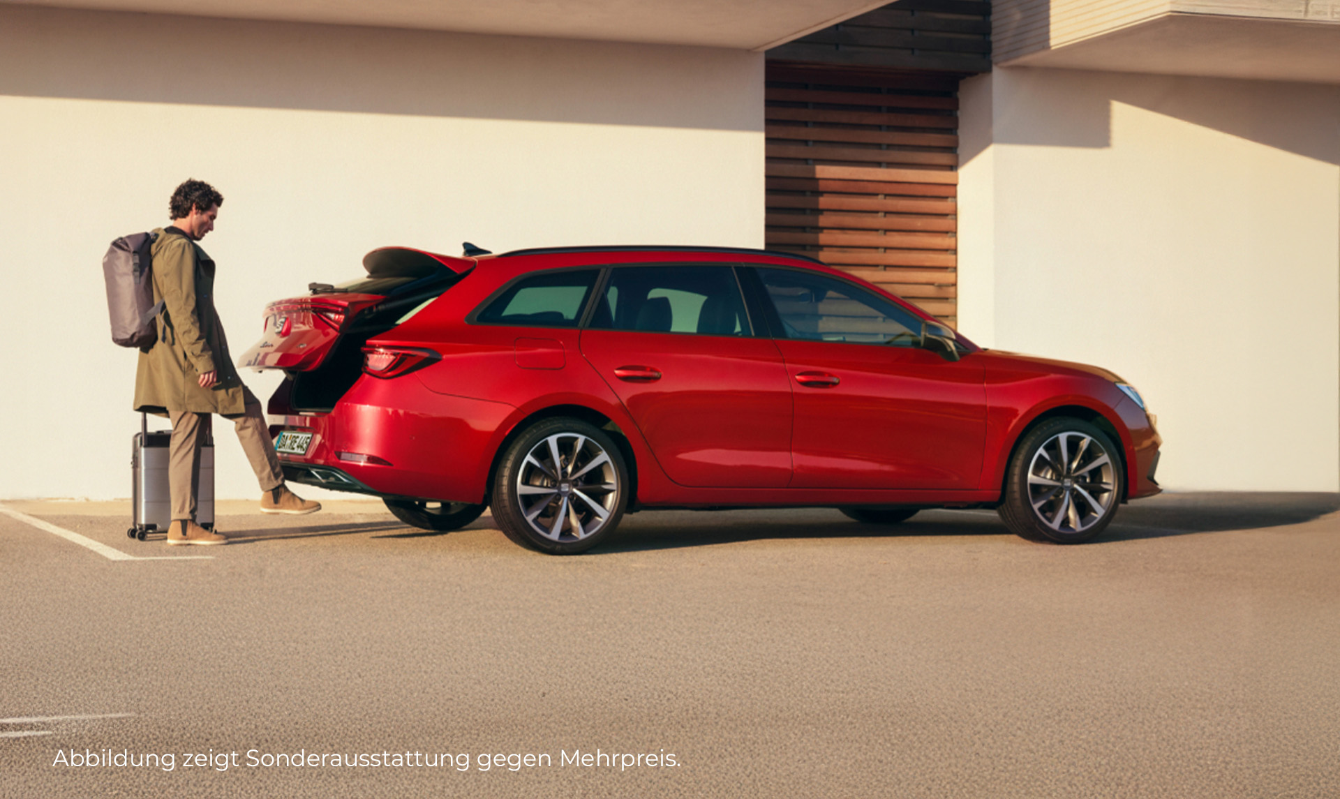 Auf dem Bild ist ein roter SEAT Leon Sportstourer von der Seite abgebildet. Die Front zeigt nach rechts. Am Kofferraum steht ein Mann mit Gepäck, der mit Hilfe der Fußsensoren am Kofferraum die Klappe öffnet. Im Hintergunfd ist ein modernes, schlichtes Gebäude.
