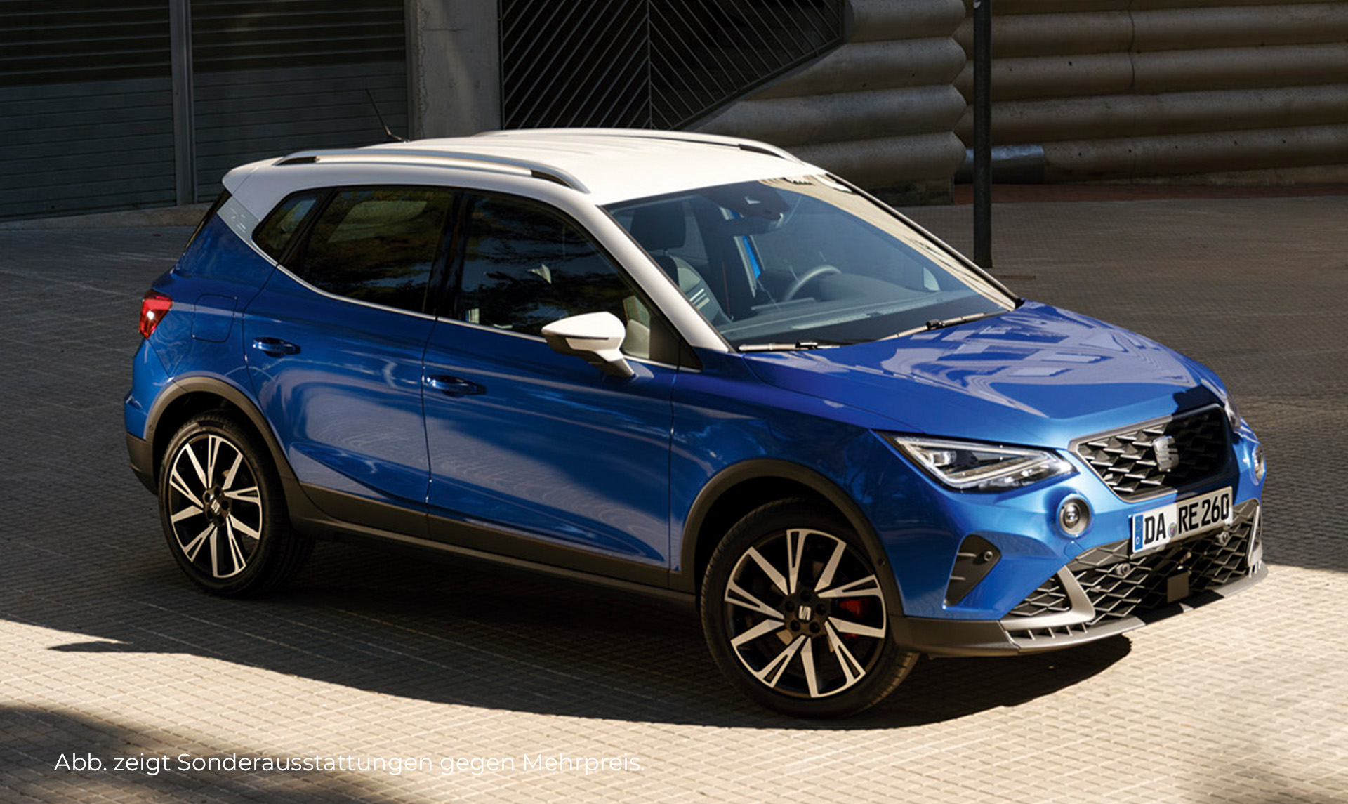 Ein SEAT Arona in blau mit weißem Dach von einer Seitenperspektive parkend vor einer grauen Halle.