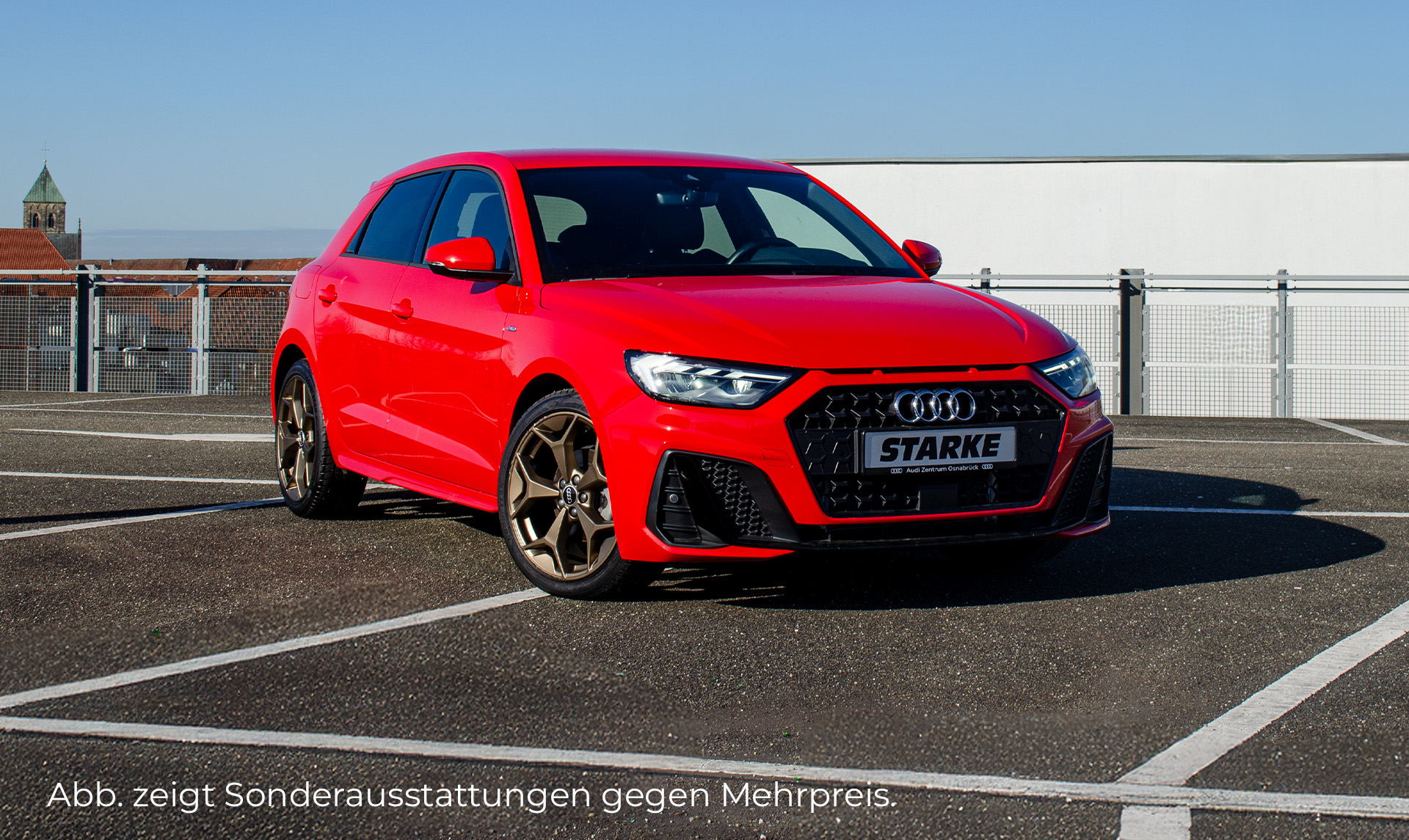Das Bild zeigt einen roten A1 auf einem Parkplatz im Hintergrund sind ein Geländer und Häuser zu sehen. Der Audi ist auf dem Bild leicht nach rechts gedreht. Zu sehen ist die Front und die linke Fahrzeugseite. 