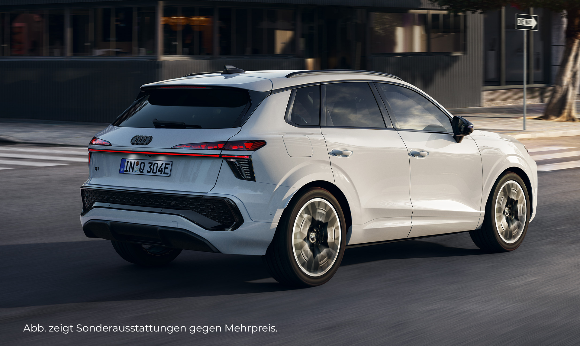Ein weißer Audi Q3 SUV Plug-in-Hybrid fährt auf einer städtischen Straße, wobei die Heckansicht im Vordergrund steht und moderne Architektur die Kulisse bildet.
