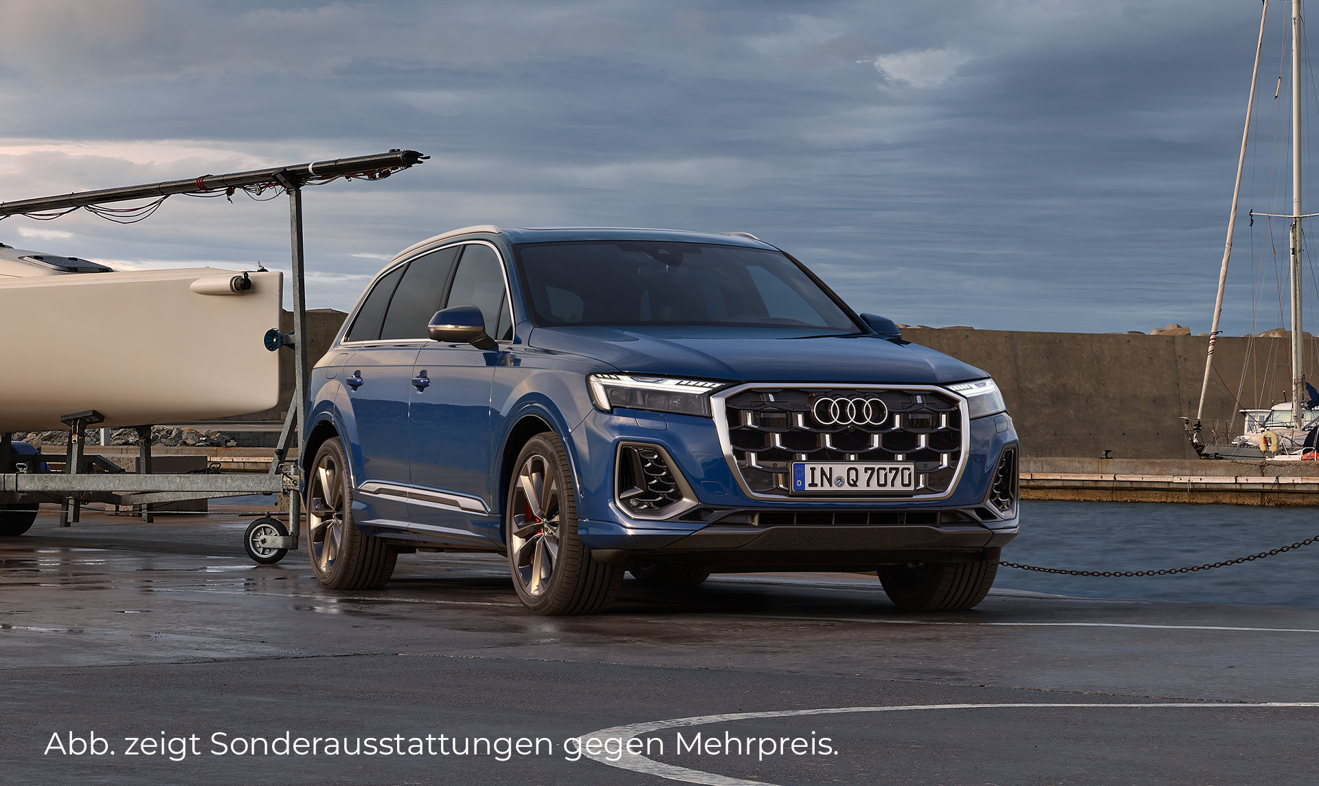 Das Bild zeigt einen blauen Q7 der auf einem Parkplatz steht. Im Hintergrund ist ein Hafen mit Schiffen. Zu sehen ist die Vorderseite und die rechte Seite vom Audi.