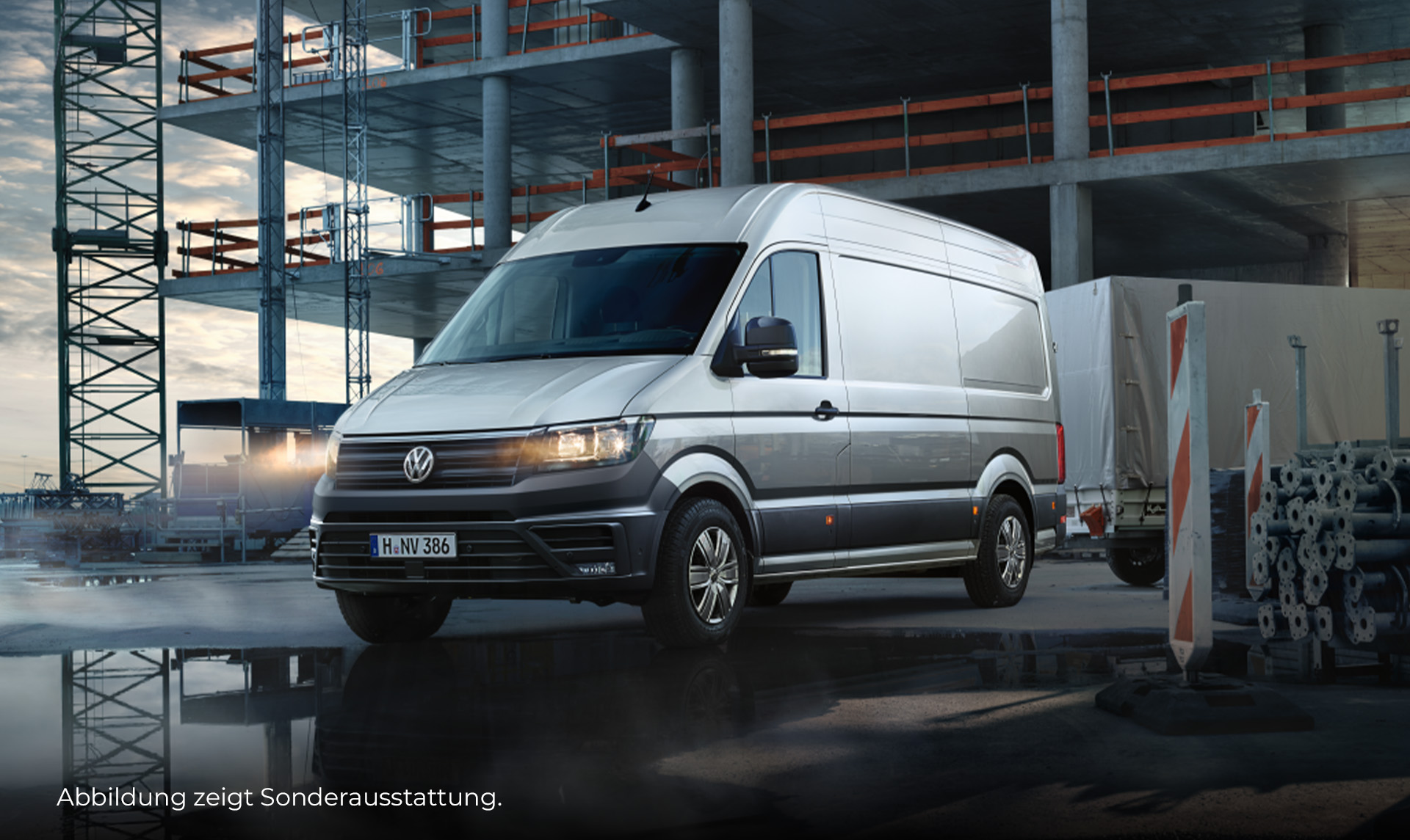 Ein silberner VW Crafter steht vor einem noch nicht vollständig errichteten Gebäude. Das Fahrzeug ist frontal und seitlich zu sehen und zieht einen geschlossenen Anhänger. Der Boden vor dem Fahrzeug ist nass, sodass sich ein Teil des Fahrzeugs und der Baustelle in der Pfütze spiegelt. Neben dem Fahrzeug stehen Baustellenabsperrungen und gestapelte Gerüststangen. Unten links im Bild steht der Hinweis: "Abbildung zeigt Sonderausstattung".