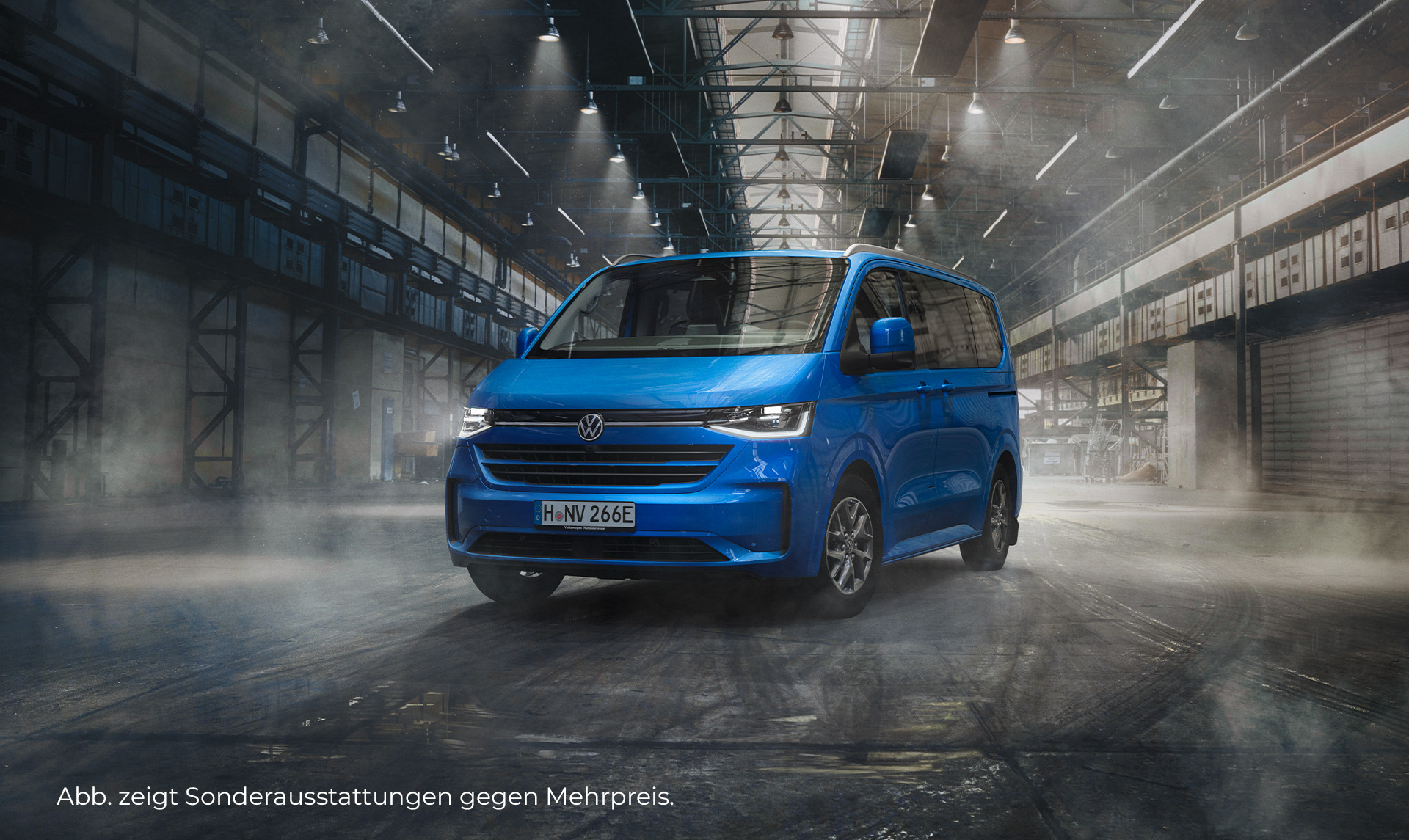 Ein blauer VW e-Caravelle steht in einer großen, beleuchteten Lagerhalle im modernen Design. Das Fahrzeug ist frontal und seitlich zu sehen. Der Hintergrund ist leicht nebelig. Unten links im Bild steht der Hinweis: "Abb. zeigt Sonderausstattungen gegen Mehrpreis".
