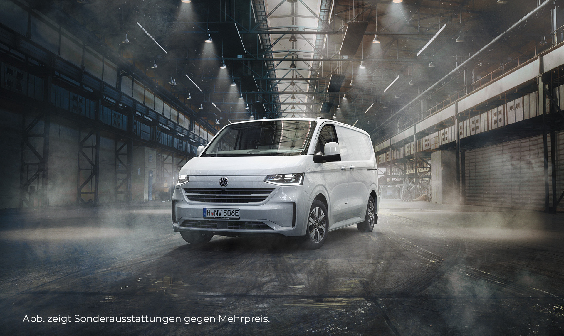 Ein weißer VW e-Transporter steht in einer großen, beleuchteten Lagerhalle im modernen Design. Das Fahrzeug ist frontal und seitlich zu sehen. Der Hintergrund ist leicht nebelig. Unten links im Bild steht der Hinweis: "Abb. zeigt Sonderausstattungen gegen Mehrpreis".