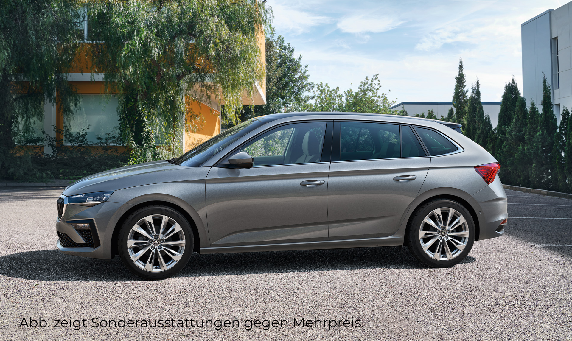 Das Bild zeigt einen seitlich stehenden grauen Skoda Scala auf einem Parkplatz. Im Hintergrund sind Bäume und drei Gebäude zu sehen.