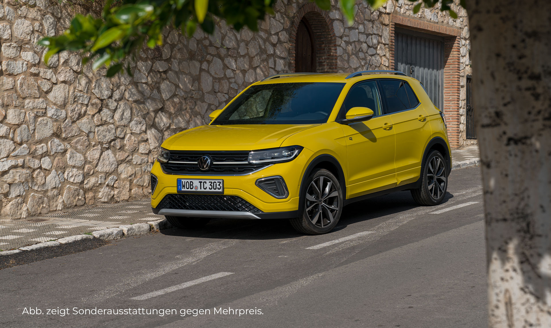 Ein gelber VW T-Cross R-Line steht auf einer Straße vor einer Steinmauer mit Holztor und Fensterläden. Das Fahrzeug ist von vorne schräg links zu sehen. Unten links im Bild steht der Hinweis: "Abbildung zeigt Sonderausstattung".