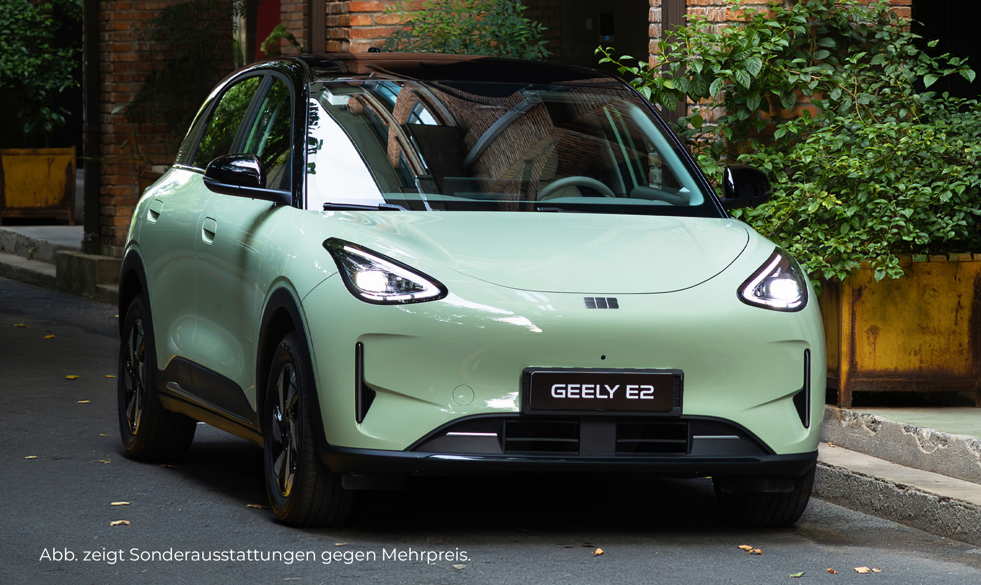 Geely E2 in hellgrüner Lackierung auf einer Straße vor einem Gebäude mit Backsteinfassade und bepflanzten Eingangsbereichen.