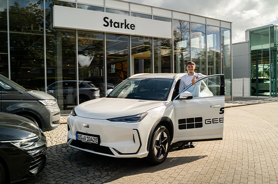 Person steht an einem Geely E5 vor dem Eingangsbereich eines Autohauses mit Geely Fahrzeug und Glasfassade im Hintergrund.