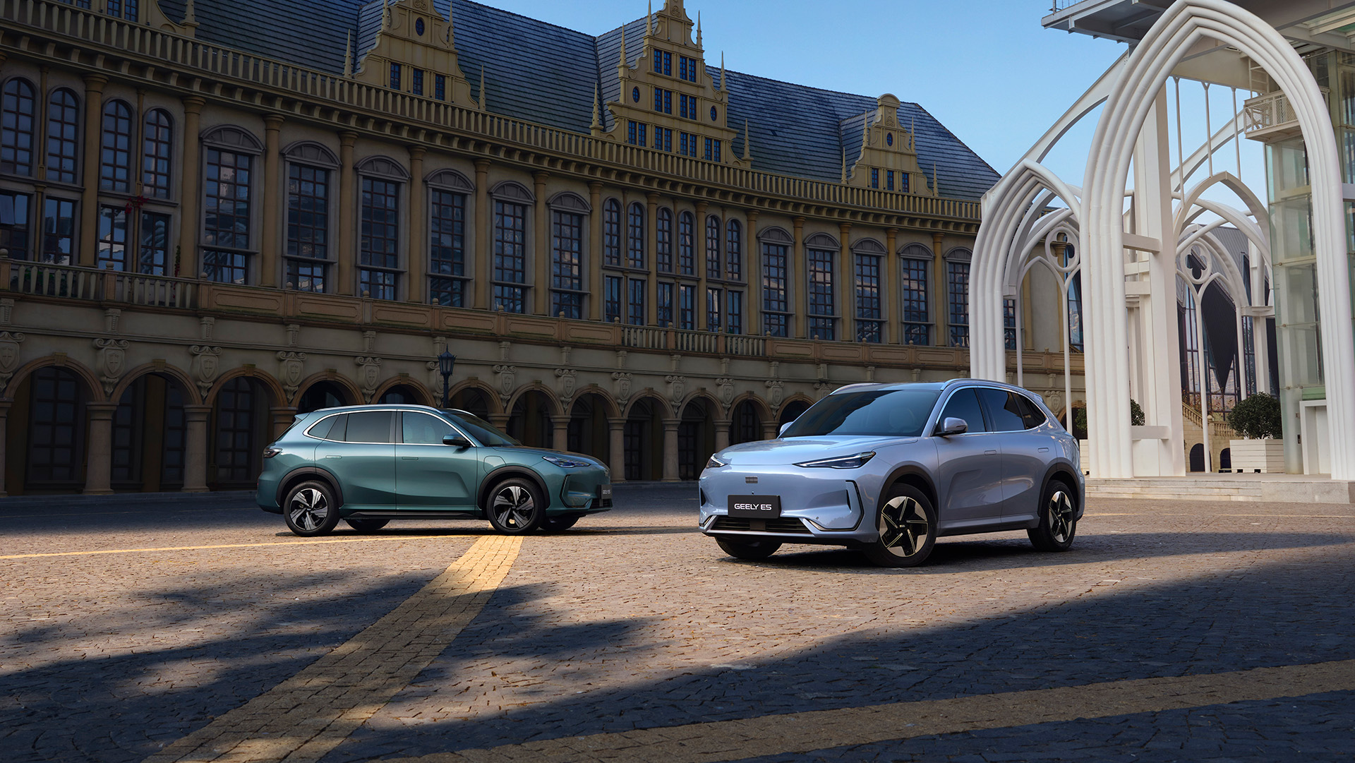 Zwei Geely E5 auf einem Kopfsteinpflasterplatz vor einem historischen Gebäude, links in Petrol in der Seitenansicht, rechts in Hellblau von vorne.