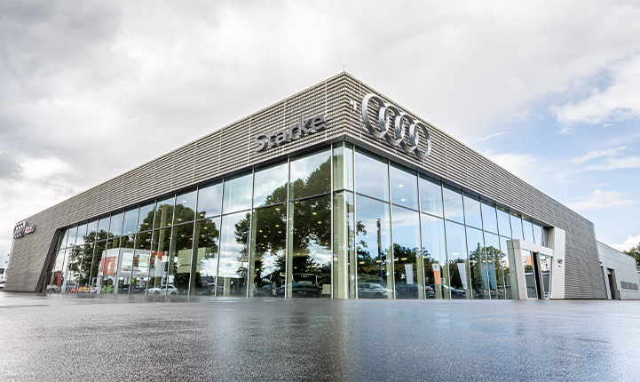 Auf dem Foto ist der Audi Vechta Standort zu sehen. Das Gebäude ist von unten Fotografiert, sodass eine Gebäudeecke in den Himmel ragt. Der Boden ist nass und der Himmel bewölkt.