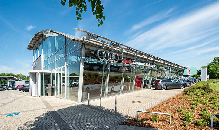 Auf dem Bild ist das Audi Zentrum Osnabrück zu sehen, der Himmel ist blau mit leichten Wolken und im unteren rechten Bild ist ein grünes Beet zu sehen. 