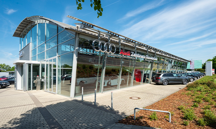 Auf dem Bild ist das Audi Zentrum Osnabrück zu sehen, der Himmel ist blau mit leichten Wolken und im unteren rechten Bild ist ein grünes Beet zu sehen. 