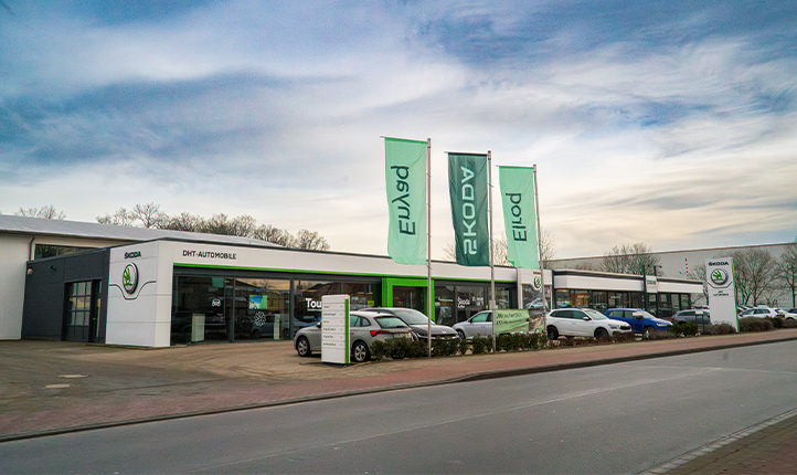 Auf dem Bild ist das DHT Automobil Gebäude zu sehen, der Himmel ist blau mit leichten Wolken. Vor dem Gebäude stehen Fahrzeuge und drei Große grüne Fahnen. Das Skoda Logo ist an einem Schild vor dem Gebäude uns links an der Wand von dem Gebäude zu sehen.